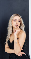 Fototapeta premium Confident woman poses indoors. A young woman with long blonde hair poses against a dark wall, showing off her stylish outfit and accessories.