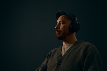 Obraz premium Portrait of a focused man with a beard wearing black over-ear headphones, listening intently against a dark, moody background. Ideal for music, podcasting, and audio technology concepts.