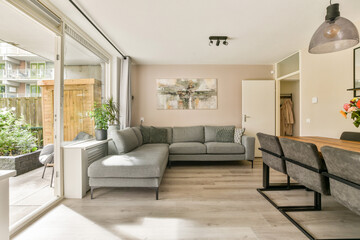 A bright, stylish modern living room featuring a gray sofa, dining table, and large windows with exposed greenery outside, enhancing a cozy atmosphere.
