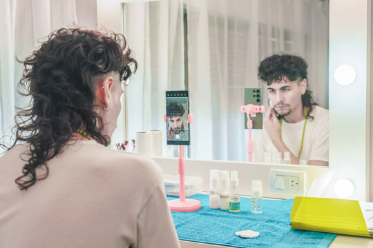 Young man filming himself applying makeup tutorial using smartphone on tripod in front of mirror - Powered by Adobe