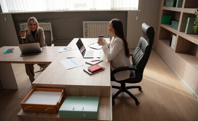 The image shows two professional women in a modern office. One is smiling and holding a coffee cup, while the other is working on a laptop. The desk is filled with documents and office supplies.