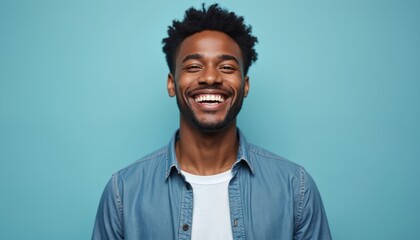 Fototapeta premium Joyful young African American man with bright smile wearing casual denim shirt and white t-shirt on plain light blue studio background. Happy face expressions and positive emotions. Success concept.