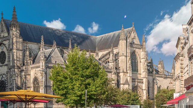 Basilique Saint-Michel timelapse in Bordeaux, France, is a gothic landmark with a tall spire. People enjoy cafes nearby, under green trees and blue cloudy skies, capturing the city's vibrant charm