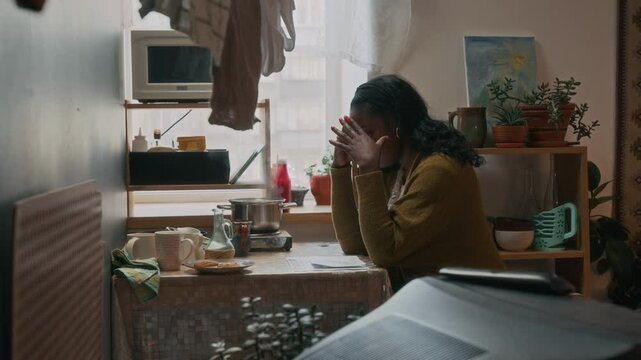 Poor African American woman sitting at lived-in kitchen table, feeling anxious, looking at past due bills