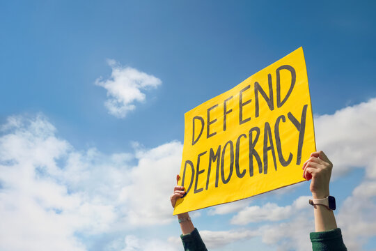 Person holding up a Protest sign that reads "Defend Democracy" against a blue cloudy sky