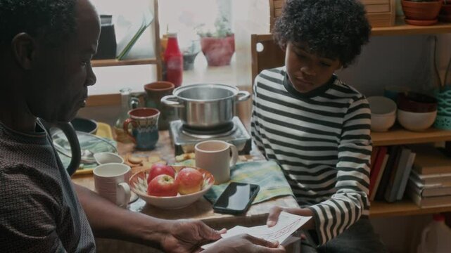 African American boy from poor family listening to his father telling about past due bills, sitting in small kitchen