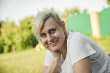 A joyful woman with short blonde hair, wearing a white t-shirt, smiles softly while resting her head on her hand. She sits in a sunny outdoor garden with green trees and a fence in the background.