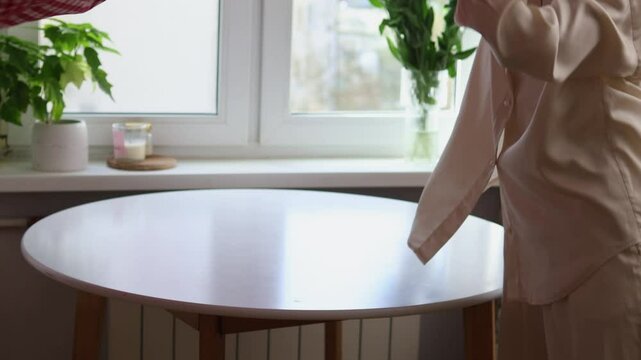 Women's hands laying a red checkered tablecloth on a white round table by the window. Preparing a festive table.