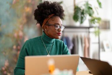 A young woman with curly hair, glasses, and a green sweater is focused on her laptop while working in a modern office space with a clothing rack and plants in the background.