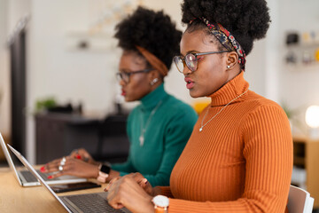 Two women with glasses and curly hair are collaborating at a wooden office table with laptops in a bright, modern workspace featuring indoor plants and stylish decor.