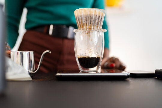 A close-up view of a pour-over coffee dripper placed on a transparent glass cup as freshly brewed coffee drips down, with a blurred background featuring a person preparing coffee.
