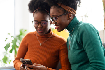 Two women sit close together, smiling as they look at a smartphone screen, sharing a moment of laughter and connection in a bright and cozy home environment.
