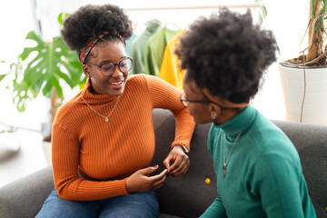 Two women sit together on a gray sofa, smiling as they chat in a cozy and stylish living space filled with natural light and a warm, inviting atmosphere.