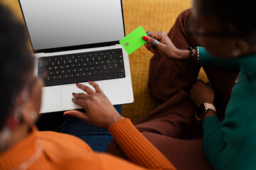 Two people sitting together on a couch using a laptop while one holds a bright green credit card, engaged in an online shopping or payment activity.