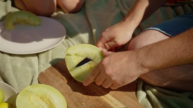 Father with daughter at picnic, man slicing melon into wedges. Dad preparing fresh fruit while child arranges pieces on plate. Hands carefully cutting juicy melon as girl helping with serving during