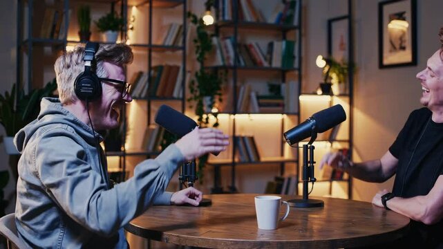 Two people recording a podcast video, sitting at a round table with microphones. Side angle captures their animated conversation and cozy studio setup.