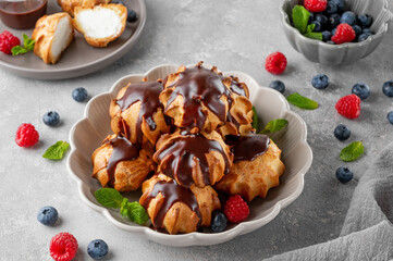 Profiteroles with vanilla cream, fresh berries and chocolate glazed in a bowl on a gray concrete background. A delicious French dessert.