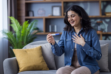 Happy young Indian woman sitting on the couch at home and rejoicing at the news of pregnancy,...