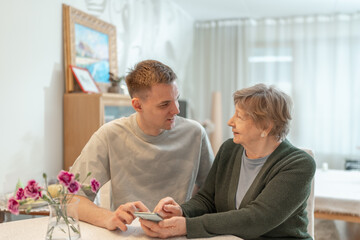 Fototapeta premium Young man helping senior woman with a smartphone at home. Concept of technology, education, and senior digital adaptation. Grandson teaches grandmother how to use digital device. Assisted living.