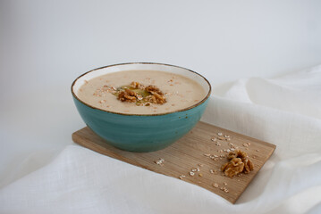 Creamy oatmeal porridge in a teal bowl on a wooden board on a white background. Healthy food with walnuts, kiwi, and oat flakes. Nutritious breakfast morning meal.