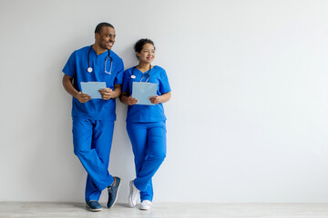 African American man and woman in medical scrubs looking aside at copy space, medical workers leaning casually against a white wall while holding clipboards, appearing cheerful and professional