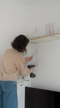 Young confident woman hanging shelf on wall. Female screws a screw into book shelf mount. creative girl makes house cozy. DIY home improvement, cozy room transformation. Vertical video