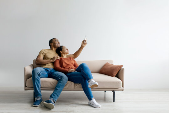 Split system. Cheerful young black couple with remote relaxing under air conditioner, sitting on couch over white studio background. Happy african american spouses using climate control