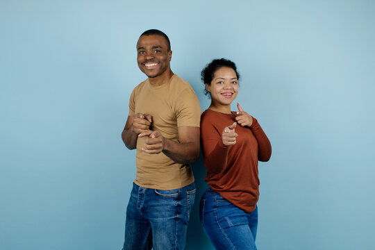 Smiling African American couple pointing confidently at the camera with hands, black young man and woman standing back to back in a casual studio setting with cheerful engaging expressions