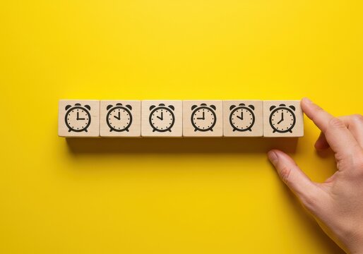 A hand picking up a wooden block with a clock icon among others on yellow background symbolizing time management