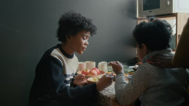 African American boy eating porridge for breakfast with his older brother, while his unrecognizable mother stroking his back