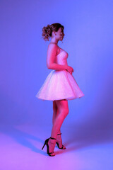 Model poses in a sparkly pink dress with heels under purple lighting in a modern studio setting