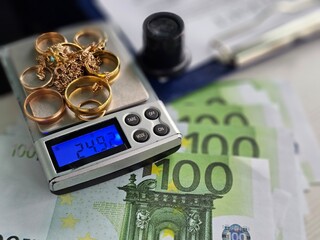 Gold rings necklaces are displayed on a digital scale with euro notes underneath, indicating a...
