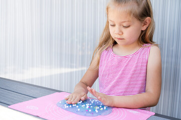Young girl in a pink striped dress playing with blue sensory slime and foam beads on a mat,...