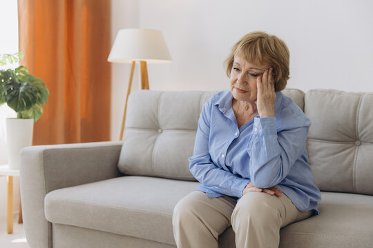 Sick older senior woman sit on sofa feel pain dizziness coping with morning headache concept, upset tired old woman touching sore head suffer from terrible migraine mental problem