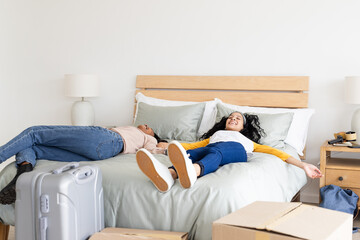 Relaxing on bed, two diverse female friends enjoying break after unpacking in new home