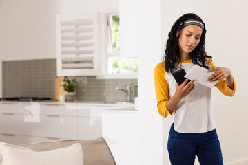 At home, woman reading mail while holding smartphone, looking focused and thoughtful