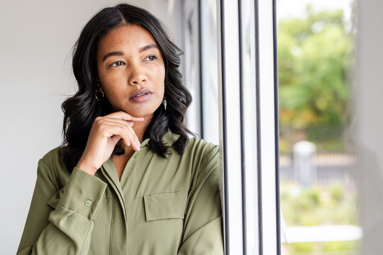 Thoughtful woman in green shirt looking out window, contemplating ideas, at home