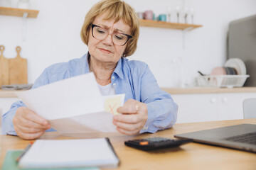 Shocked puzzled senior woman reading paper document, reviewing agreement terms, checking bills for...