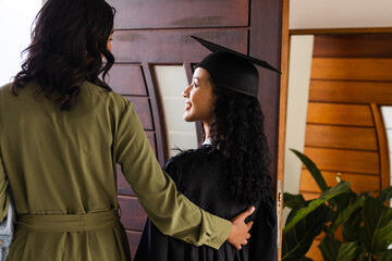 Proud mother embracing daughter in graduation gown at home, celebrating achievement