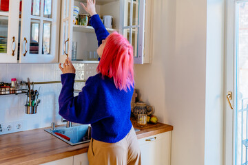 Young woman with bright pink hair reaching up into kitchen cabinet for cup, casual daily routine...