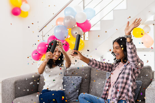 Celebrating at home, two diverse female friends joyfully popping confetti with balloons around