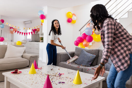 Cleaning up after party, two diverse female friends laughing and sweeping confetti, at home