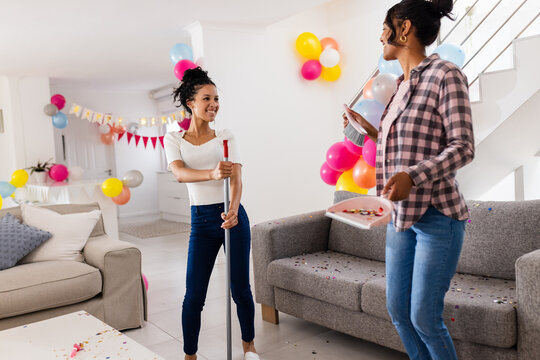 Diverse female friends cleaning living room after party, smiling and holding mop and tray, at home - Powered by Adobe