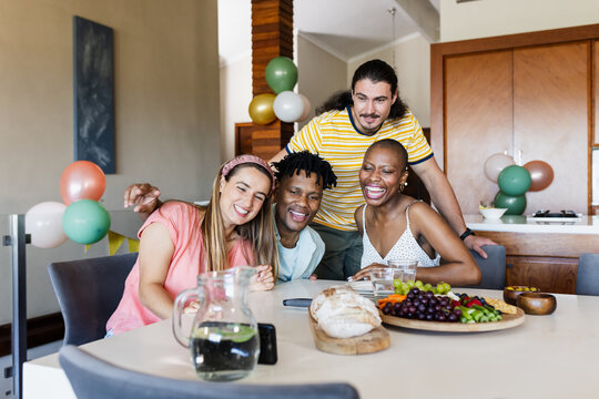 Smiling diverse friends enjoying snacks and drinks at home, celebrating together