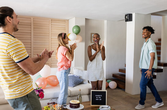 Diverse friends celebrating new job with cake and balloons in living room, smiling joyfully, at home