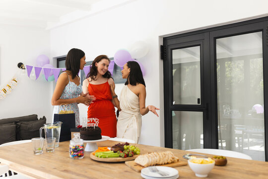 Celebrating birthday, three diverse female friends chatting and laughing at home with snacks
