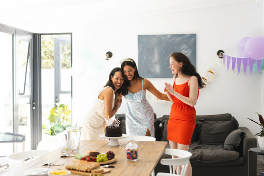 Celebrating birthday at home, three diverse female friends laughing near cake and decorations