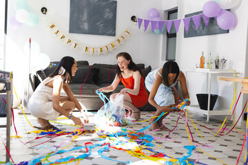 Diverse female friends celebrating birthday, cleaning colorful streamers in living room, at home