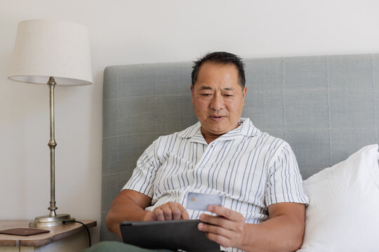 Asian man using tablet and credit card while relaxing on bed at home