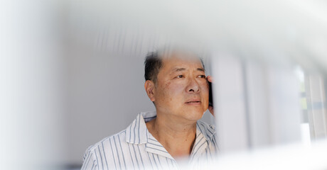 Asian man in pajamas looking out window, appearing thoughtful and reflective
